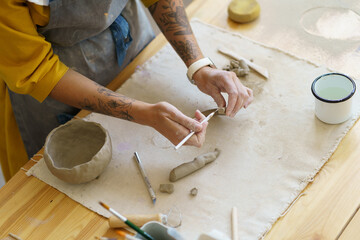 Cropped shot of woman working with clay in pottery studio during masterclass, learning basic hand-building techniques during ceramics workshop, female potter in apron at workplace. Art therapy concept