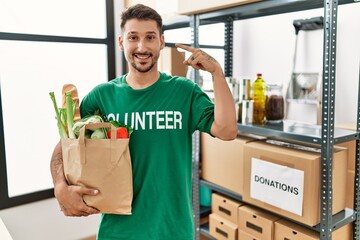 Canvas Print - Young hispanic man wearing volunteer t shirt holding groceries pointing finger to one self smiling happy and proud