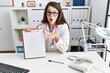 © Krakenimages.com - Young doctor woman holding clipboard at the clinic scared and amazed with open mouth for surprise, disbelief face