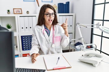 Wall Mural - Young doctor woman wearing doctor uniform and stethoscope at the clinic pointing finger up with successful idea. exited and happy. number one.