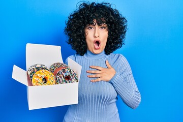 Poster - Young middle east woman holding tasty colorful doughnuts box scared and amazed with open mouth for surprise, disbelief face