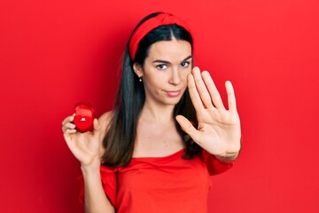 Young brunette woman holding engagement ring for proposal with open hand doing stop sign with serious and confident expression, defense gesture