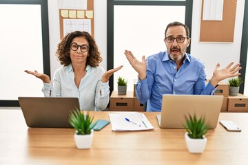 Wall Mural - Middle age hispanic woman and man sitting with laptop at the office clueless and confused expression with arms and hands raised. doubt concept.
