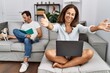 © Krakenimages.com - Hispanic middle age couple at home, woman using laptop looking at the camera smiling with open arms for hug. cheerful expression embracing happiness.