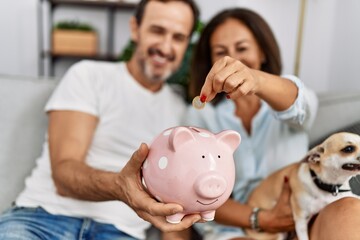 Sticker - Middle age man and woman couple holding piggy bank sitting on sofa with dog at home