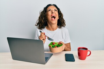 Sticker - Beautiful middle age woman working at the office eating healthy salad angry and mad screaming frustrated and furious, shouting with anger looking up.