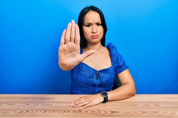 Beautiful hispanic woman with nose piercing sitting on the table doing stop sing with palm of the hand. warning expression with negative and serious gesture on the face.