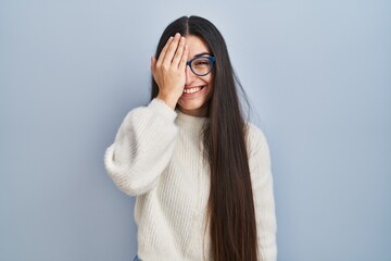 Poster - Young hispanic woman wearing casual sweater over blue background covering one eye with hand, confident smile on face and surprise emotion.