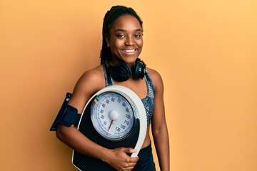 Poster - African american woman with braided hair wearing sportswear holding weighing machine looking positive and happy standing and smiling with a confident smile showing teeth