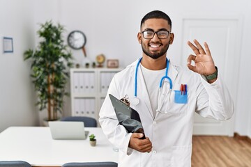 Poster - Young indian man wearing doctor uniform and stethoscope smiling positive doing ok sign with hand and fingers. successful expression.