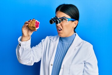 Poster - Beautiful brunette jeweller woman holding geode stone wearing magnifier glasses in shock face, looking skeptical and sarcastic, surprised with open mouth