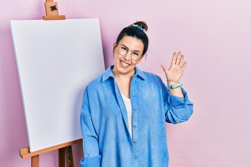 Poster - Young hispanic woman standing by painter easel stand waiving saying hello happy and smiling, friendly welcome gesture