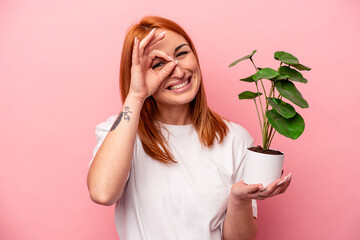 Wall Mural - Young caucasian woman holding a plant isolated on pink background Young caucasian woman holding a plant isolated on pink background excited keeping ok gesture on eye.