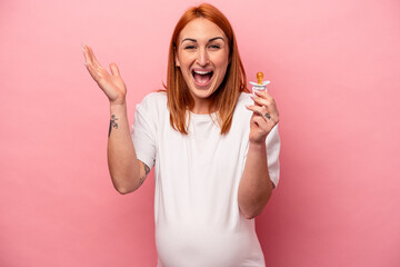 Wall Mural - Young caucasian pregnant woman holding pacifier isolated on pink background receiving a pleasant surprise, excited and raising hands.