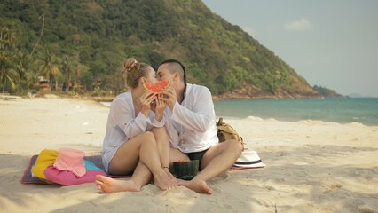 The cheerful love couple holding and eating slices of watermelon on tropical sand beach sea. Romantic lovers two people caucasian spend summer weekend in outdoor. Hat, backpack white shirt beachwear.