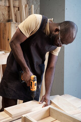 Wall Mural - Vertical portrait of black carpenter using power drill while building wooden furniture in workshop