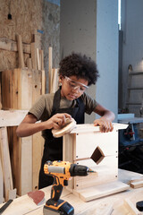 Wall Mural - Vertical portrait of young black boy building wooden birdhouse in workshop and sanding in carefully