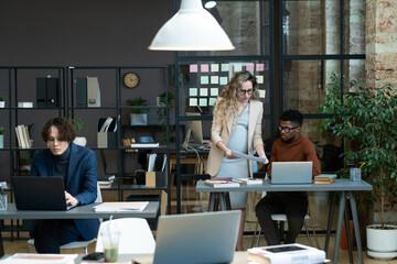 Wall Mural - Young pregnant businesswoman showing the document to her colleague while they working at office on computers with other people