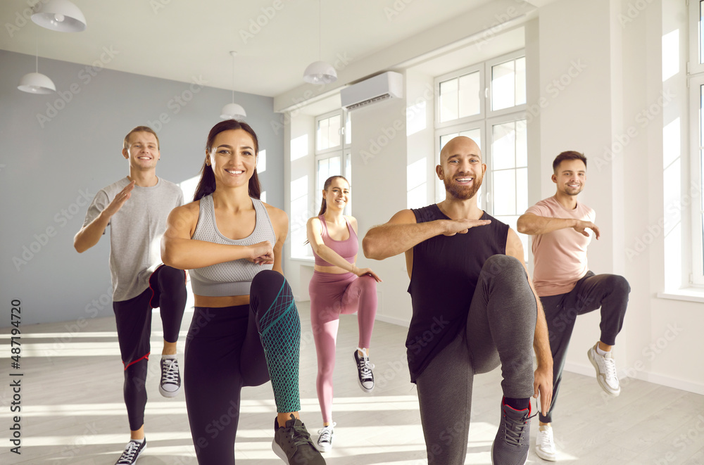 Group of young sporty people doing exercises together in bright and spacious gym. Happy men and women doing aerobics exercises lifting high knees in gym. Fitness sport aerobics and people concept. Sto...