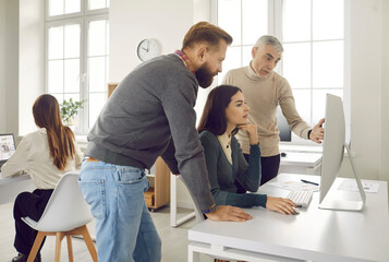 Group of three people looking at computer screen and discussing something. Team of young and older corporate employees working on online project in modern light company office interior