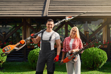 Wall Mural - Portrait of happy caucasian man and woman posing at beautiful garden with electric hedge trimmers in hands. Male and female gardeners enjoying seasonal work with plants.