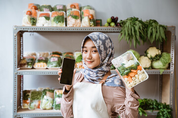 Wall Mural - Woman greengrocer showing vegetables and showing a phone screen