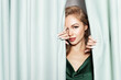 Sensual pretty young blond girl wearing celebratory evening dress, with perfect makeup peek out green curtains. Alluring caucasian female posing near curtain. Festivity, skincare cosmetics advertising