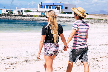 Wall Mural - Young tourists couple wakl holding hands in back view at the beach. Summer tourism holiday vacation in tropical resort scenic place. Young man and woman enjoy sun and sea leisure