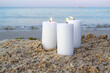 Three large white paraffin candles burn on sand beach background sea waves. Decorative Burning candles on sea coast close up. Candlelight, candle flame. Romantic mood backdrop. Concept date relaxation