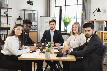 Wall Mural - Team of five multiracial business people in formal clothes sitting together at office desk, smiling and looking at camera. Conference meeting of diverse colleagues.