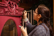 Woman aging wooden cupboard with little brush with brilliant carved ornaments of bright red. Scrupulous process of patinating furniture. Eco-friendly re-usage. Old things getting new life.
