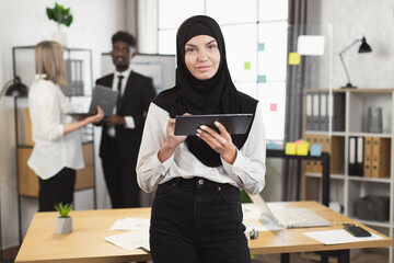 Wall Mural - Confident muslim woman in hijab looking at camera while posing at office with clipboard in hands. African american and caucasian colleagues having discussing on background.