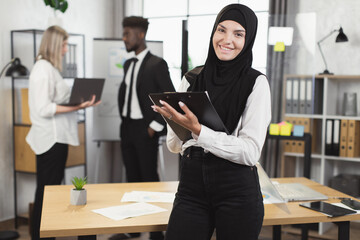 Wall Mural - Muslim businesswoman with clipboard in hands smiling and looking at camera while her multiracial colleagues talking on background. Meeting at office of successful partners.