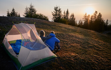 Wall Mural - Back view of two travelers sitting on grass in front of tent set up on mountain hill, early in the morning and contemplating on sunrise. Soft sun rays lighting campsite.