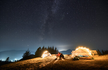 Wall Mural - Girl hugging her boyfriend, they sitting together on grass between two tents iluminated by light bulbs and resting under beautiful starfull sky at night in the mountain hills.