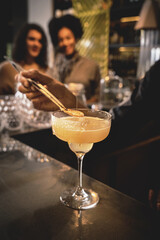 Wall Mural - Bartender garnishing a Margarita cocktail - blurry cheerful girls party in the background smiling together.