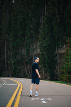A Young Man In Dark Clothes Walks On A Footpath In The Mountains Against The Backdrop Of Coniferous Trees And Looks Away. Vertical