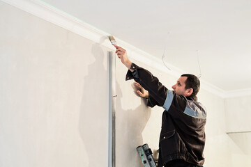 Man paints a plaster ceiling plinth with a brush. Current repairs in the apartment. Restoration indoors. Plasterboard panel.