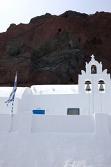 Wall Mural - View of a white Greek Orthodox Church with a greek flag and the volcanic landscape of the famous red beach in the background