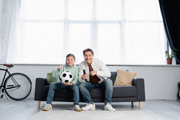 Father and son with remote controller and football ball watching match at home