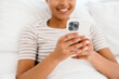 © Drobot Dean - Young black woman smiling and using cellphone while sitting in bed