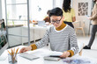 © AnnaStills - Asian serious businesswoman in eyeglasses sitting at the table and typing document on computer at office with her colleagues in the background