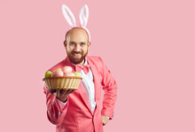 Portrait Of Happy Funny Handsome Bearded Young Man In Pink Suit, Bow Tie And Cute Bunny Ears Standing Isolated On Color Background, Looking At Camera, Showing Wicker Bowl With Easter Eggs And Smiling