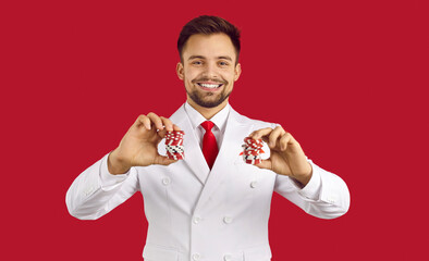 Portrait of smiling confident poker professional. Happy lucky handsome young man in elegant white suit with neck tie standing isolated on red background and holding two stacks of casino chips in hands