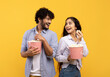 Overjoyed indian man and woman eating popcorn, standing back to back and smiling at each other, yellow background