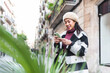 © dzmatph - Positive Chinese female in outerwear text messaging on modern cellphone while standing on street near blurred green plants in city