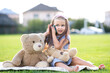 © bilanol - Pretty little child girl sitting in summer park with her teddy bear toy talking on mobile phone smiling happily outdoors in summer.