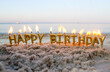 Candles Gold Letters Happy Birthday Burning on Background of Blurry Sea Waves on Sandy Beach. Burning Candles for Cake on Seaside. Concept Festive, Celebrating, Party, Birthday, Birthday Celebration