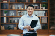 © Liubomir - Portrait of a successful Asian teacher, a man in a shirt looking at the camera and smiling, in the classic office of the university director