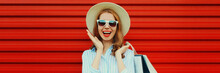 Portrait Of Happy Surprised Laughing Woman With Shopping Bags Wearing Summer Straw Hat And Shirt On Colorful Red Background, Blank Copy Space For Advertising Text